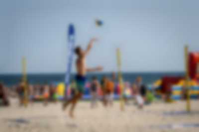 chłopak w podskoku zagrywający piłkę podczas gry w siatkówkę plażową. w tle plażowicze i morze