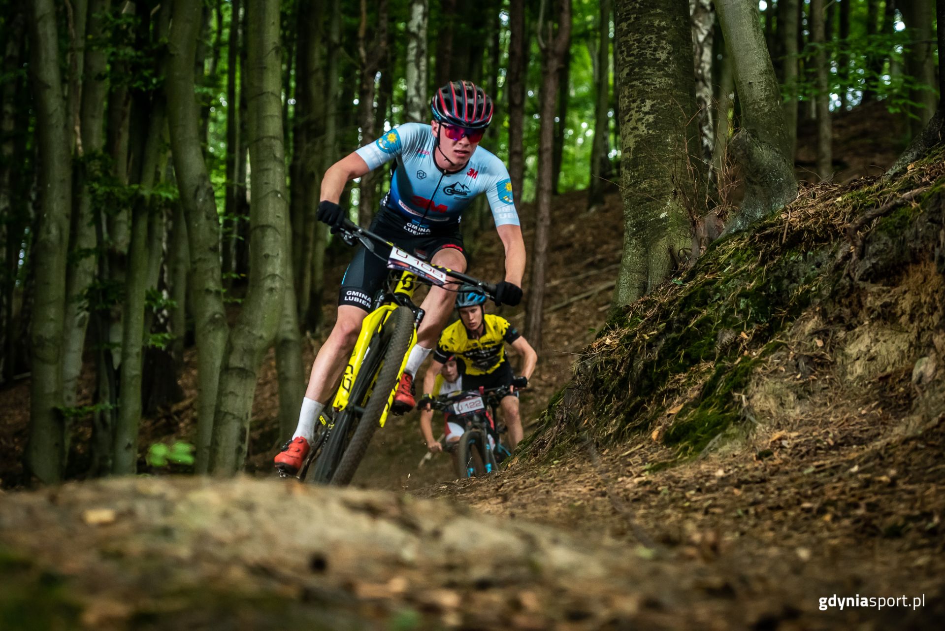 Kolarz MTB na żółtym rowerze w lesie ścigający się w zawodach. za nim kolejni 2 ścigający go zawodnicy